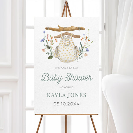 Printed foam core baby shower welcome sign featuring a soft watercolor baby romper illustration with rustic wooden hanger and floral elements. Professionally printed on thick foam board for a durable, high-quality finish. Available in multiple sizes to suit various event spaces. Perfect for welcoming guests to a boho, woodland, or gender-neutral baby shower. Easy to display on an easel or table, making it a great addition to your baby shower decor.