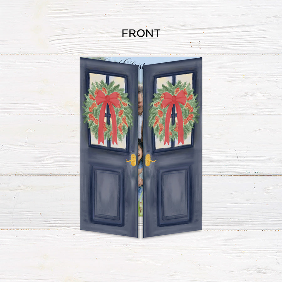 Personalized Doorway to Christmas gatefold card featuring navy blue doors decorated with festive evergreen wreaths, red bows, and elegant gold Merry Christmas lettering. The doors open to reveal a family photograph with a customizable holiday greeting and family name. - front folded.