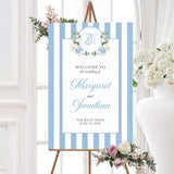 Wedding welcome sign featuring powder blue and white vertical stripes, floral crest with hydrangeas and white roses, and elegant fonts, ideal for classic spring wedding ceremonies.