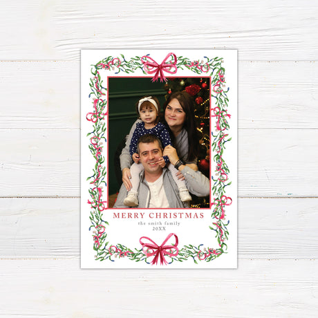 Personalized Christmas photo card featuring a family portrait framed by watercolor mistletoe, greenery, berries, and ribbon bows. The back features a coordinating watercolor bow.