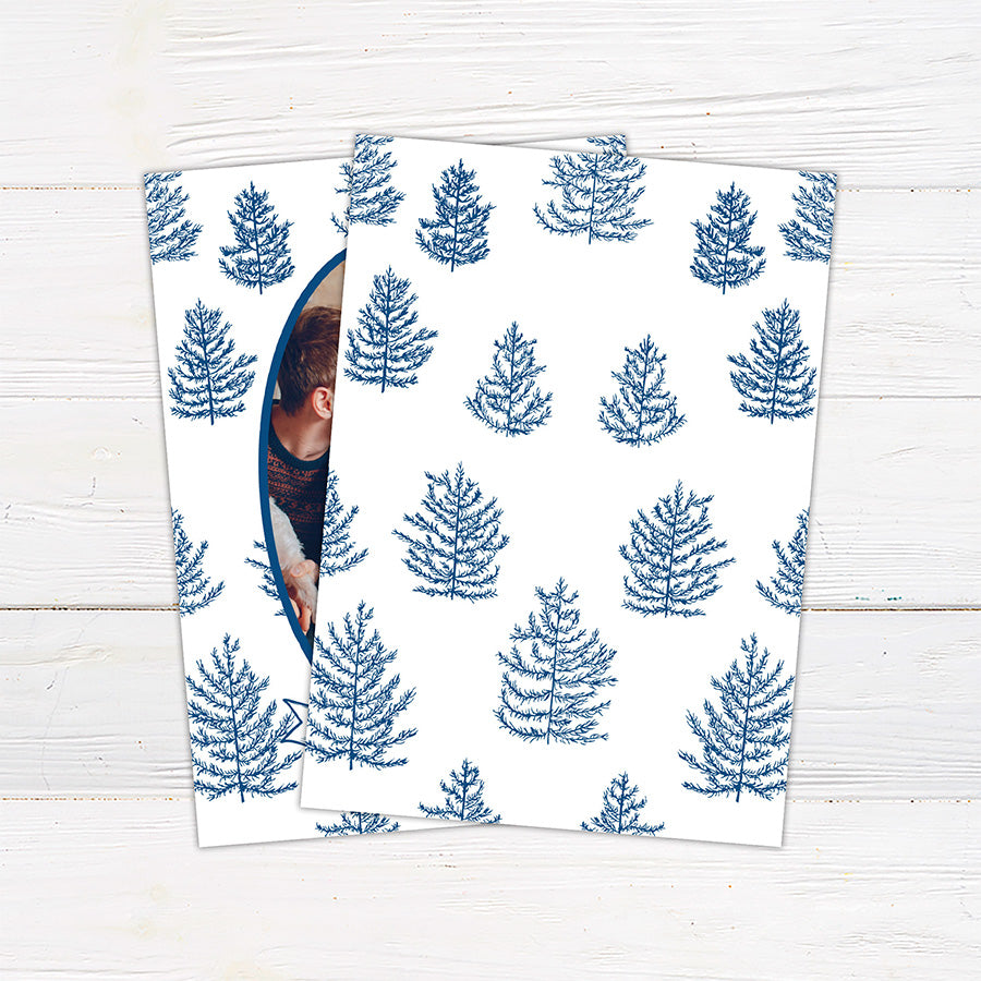 Personalized family Christmas photo card featuring an oval portrait surrounded by hand-drawn evergreen pine trees. A ribbon banner displays a Merry Christmas greeting and custom family name. blue