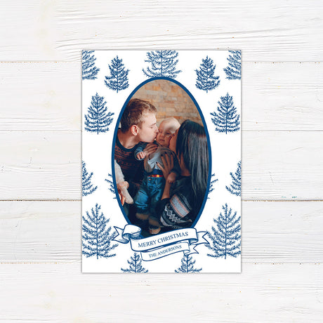 Personalized family Christmas photo card featuring an oval portrait surrounded by hand-drawn evergreen pine trees. A ribbon banner displays a Merry Christmas greeting and custom family name. blue