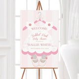 A tickled pink baby shower welcome sign shaped like a dress with ballet shoes. The design features soft floral accents and light pink stripes.